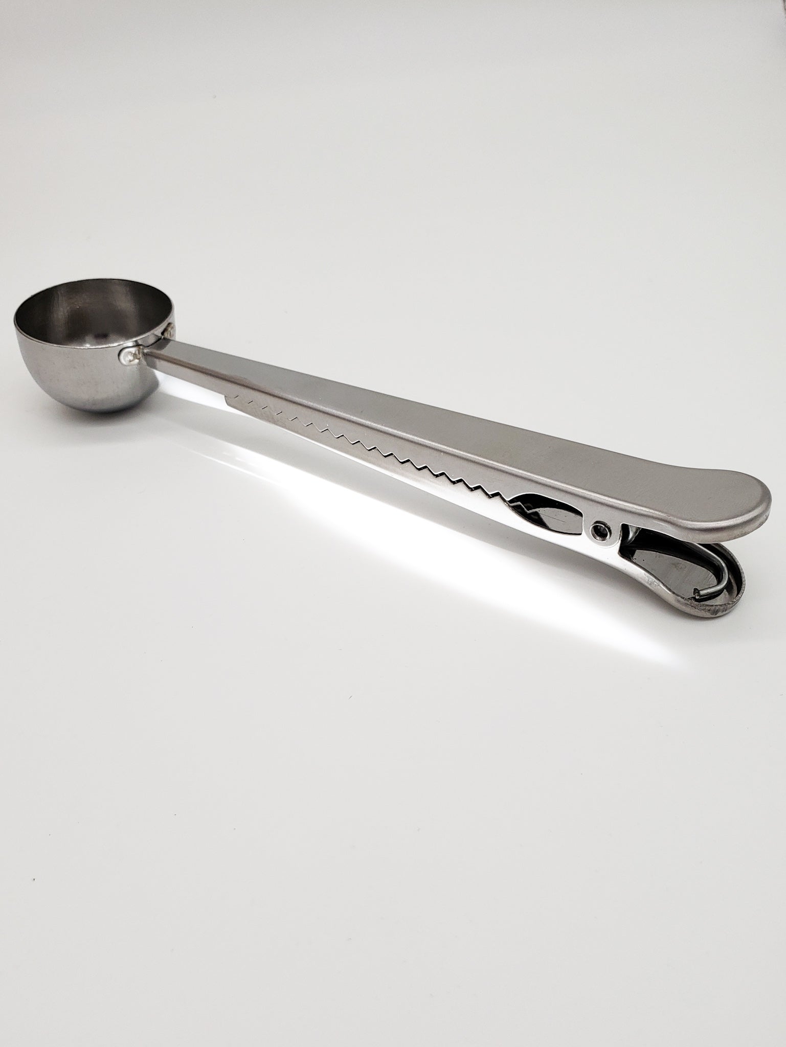 Coffee gator clip spoon. A metallic spoon with an approximately 7 inch handle to reach down into coffee package.  The spoon is very deep and full round to easily scoop coffee and the handle is a clip with gator teeth to help close and clip your coffee package closed for freshness and convenience.